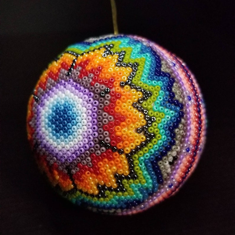 May include: A spherical ornament, intricately beaded with a rainbow of colours. The design features a geometric pattern with orange, blue, green, and purple beads. The craftsmanship suggests a handmade item, suitable for festive decorations. The diameter is approximately 7 cm.