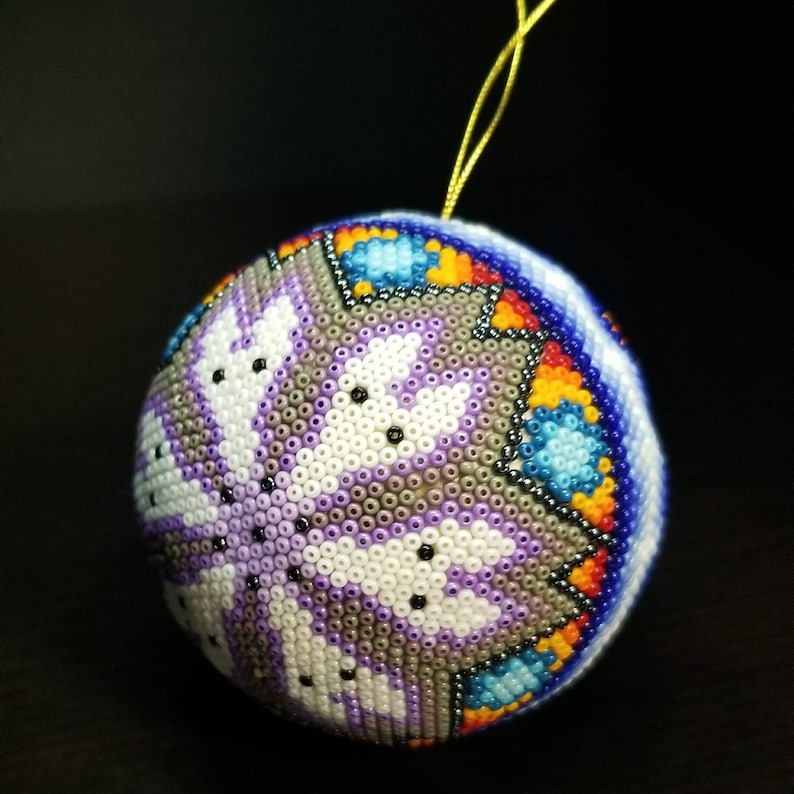 May include: A spherical Christmas bauble, intricately beaded with a geometric pattern. The design incorporates white, purple, grey, blue, orange, and black beads. A gold hanging string is attached. The bauble is a festive decoration.