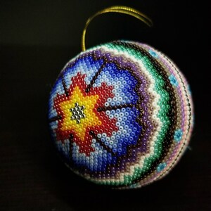 May include: A beaded bauble with a gold hanging loop. The sphere is covered in small, colourful beads in a floral pattern. The design features a central yellow flower with red and blue accents, surrounded by bands of green, purple, and white.