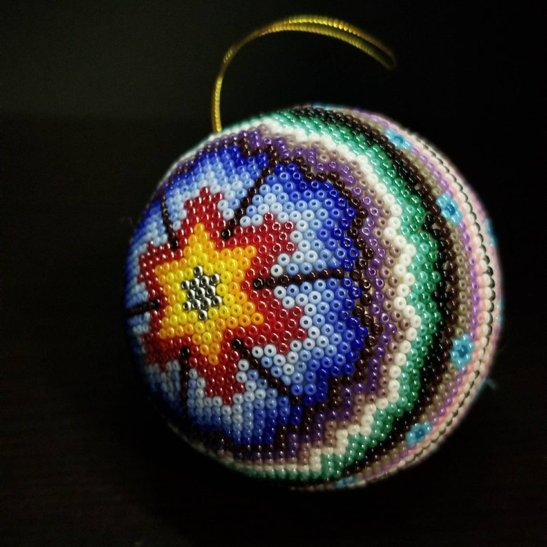 May include: A beaded bauble with a gold hanging loop. The sphere is covered in small, colourful beads in a floral pattern. The design features a central yellow flower with red and blue accents, surrounded by bands of green, purple, and white.