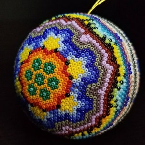May include: A spherical ornament intricately decorated with small, colourful beads. The design features concentric circles of blue, yellow, orange, and green. A gold string is attached for hanging. The ornament is likely handmade, with a diameter of approximately 8 cm.