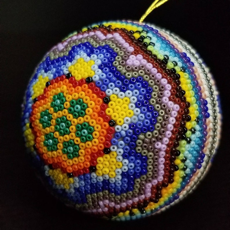 May include: A spherical ornament intricately decorated with small, colourful beads. The design features concentric circles of blue, yellow, orange, and green. A gold string is attached for hanging. The ornament is likely handmade, with a diameter of approximately 8 cm.