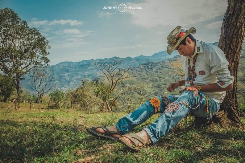 Peut inclure: Une personne portant un chapeau de paille est assise sur une colline herbeuse, regardant ses mains. Elle porte une chemise blanche avec des broderies color&eacute;es et un jean bleu avec des broderies color&eacute;es. La personne est assise devant un grand arbre avec un fond vert et brun.