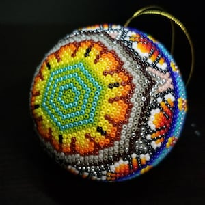 May include: A spherical, beaded decoration with a detailed design. The ornament displays a central pattern of turquoise, yellow, and orange beads, encircled by a mosaic of white, silver, and other colourful beads. A gold string is attached for hanging.