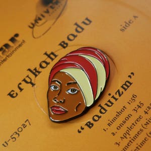 May include: A close-up of a vinyl record with a gold background and black text. The text reads "Kedar entertainment Erykah Badu" and "U-53027". The record also has a red and white enamel pin of Erykah Badu on it. The pin is a profile of Erykah Badu with a red and white turban on her head.
