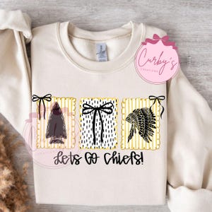 Op de afbeelding: Crèmekleurige sweatshirt met drie grafische ontwerpen: een pijl, een vlinderdas en een Indiaanse hoofdtooi, allemaal in zwart en wit. De tekst "Lets Go Chiefs!" is onder de ontwerpen gedrukt.