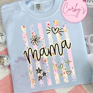 May include: Light blue t-shirt with the word "mama" in a black script font. Vertical stripes with floral patterns and decorative elements like hearts and stars are featured on the shirt. The shirt has a crew neck and short sleeves.