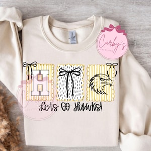 May include: Cream-colored sweatshirt with a graphic design featuring the letters "H T" and an eagle head, all in black and gold. The text "Lets Go Hawks!" is below the letters. The design includes decorative bows.