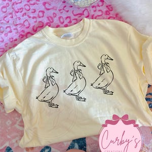 May include: A yellow t-shirt with a black graphic of three ducks wearing bows. The text "Curby's Creations" is printed on the bottom of the shirt.