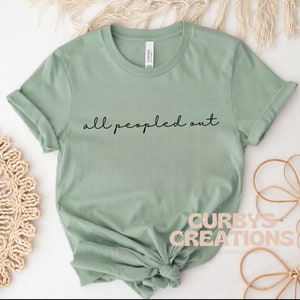 May include: A sage green t-shirt with the text "all peopled out" in black script.