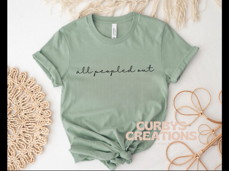 May include: A sage green t-shirt with the text "all peopled out" in black script.