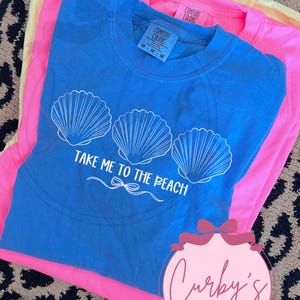 May include: A blue t-shirt with a white graphic of three seashells and the text "TAKE ME TO THE BEACH". The shirt is folded on top of a pink t-shirt.