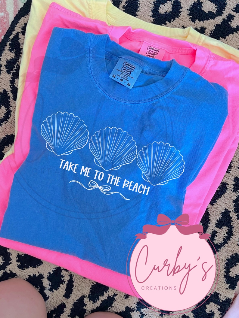 May include: A blue t-shirt with a white graphic of three seashells and the text "TAKE ME TO THE BEACH". The shirt is folded on top of a pink t-shirt.