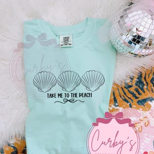 May include: A light blue t-shirt with a graphic of three seashells and the text "Take Me To The Beach".