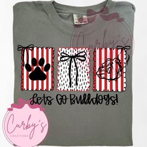 Könnte beinhalten: Graues T-Shirt mit einem grafischen Design. Es zeigt einen Pfotenabdruck, eine Schleife und einen Bulldoggenkopf, jeweils in einem rot-weiß gestreiften Rahmen. Der Text "Lets Go Bulldogs!" ist darunter gedruckt.
