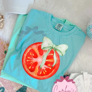 May include: A light blue Comfort Colors t-shirt featuring a watercolor illustration of a tomato slice with a green bow. A white trucker hat with "KISS ME COWBOY" in pink lettering is visible. The shirt is part of a clothing collection.
