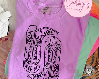 May include: A purple t-shirt with a black outline of a pair of cowboy boots. The shirt is made by Comfort Colors and has a tag that says "Curby's Creations".