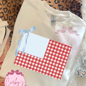 May include: White t-shirt featuring a graphic design of a blue and white gingham pattern and a red and white gingham pattern, with a blue ribbon. The shirt also has the text "Curby's Creations" in a pink circle.