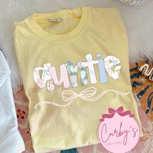May include: A yellow t-shirt with the word "auntie" in colorful patchwork letters with bows. The word is outlined in black and has a pink bow underneath. The shirt has a pink and white logo that says "Curby's Creations" in the bottom right corner.