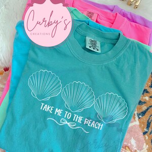 May include: A turquoise blue t-shirt with a white graphic of three seashells and the text "Take Me To The Beach".