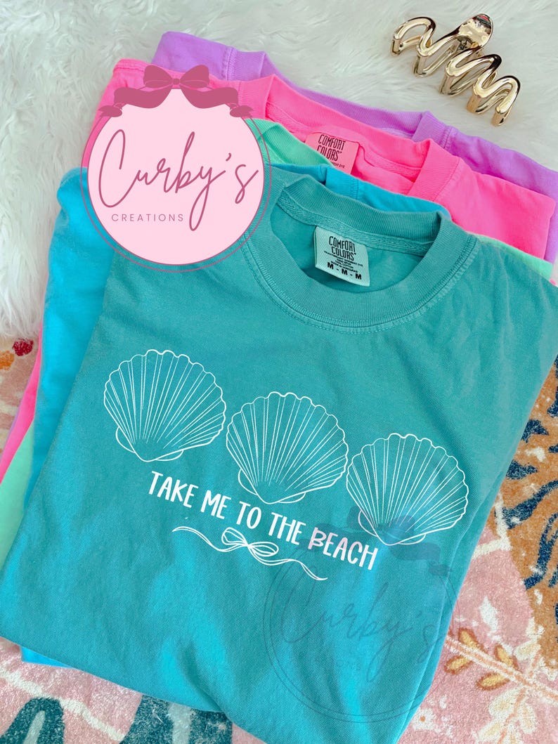 May include: A turquoise blue t-shirt with a white graphic of three seashells and the text "Take Me To The Beach".