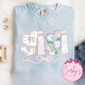 May include: Light blue t-shirt with the word "Gigi" in a patchwork style with floral and gingham patterns. The word is outlined in black and has a pink bow underneath. The text "Curby's Creations" is in a pink circle in the bottom right corner.