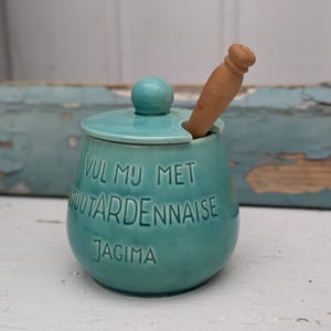 Puede incluir: Un tarro de cerámica turquesa con tapa y cuchara de madera. El tarro tiene el texto "VUL MU MET MOUTARDENNAISE JAGIMA" en relieve en la parte delantera. El tarro es redondo con un pequeño pomo en la tapa.