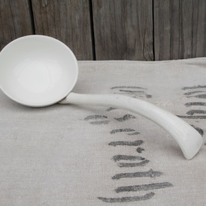 Puede incluir: Un cucharón de cerámica blanca con un mango largo. El cucharón está apoyado sobre un mantel de lino blanco con un patrón negro.