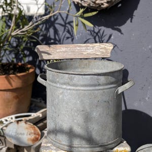 Op de afbeelding: Een gegalvaniseerde metalen emmer met twee handvatten staat op een verweerde, geverfde stoel. De emmer is cilindrisch en heeft een licht verouderd uiterlijk. Een terracotta pot en een decoratieve krans staan op de achtergrond.