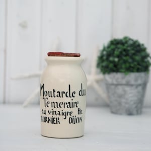 Könnte beinhalten: Weiße Keramik-Senfdose mit rotem Deckel. Die Dose trägt einen schwarzen Schriftzug mit der Aufschrift "Moutarde du le mercaire au vinaigre fin BORNIER DIJON".