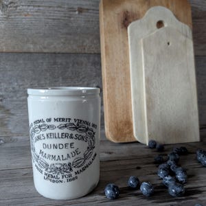 Può includere: Vaso di ceramica bianco con un'etichetta in bianco e nero che recita "MEDAL OF MERIT VIENNA 1873 JAMES KEILLER & SONS DUNDEE MARMALADE PRIZE MEDAL FOR MARMALADE LONDON 1882". Il vaso è appoggiato su una superficie di legno con mirtilli sparsi intorno.