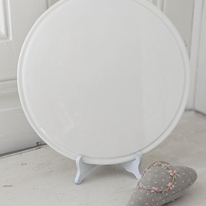 May include: A large, round, white ceramic plate on a white stand. A grey heart-shaped cushion with white polka dots and a floral wreath is in front of the plate. The plate and stand are on a white surface.