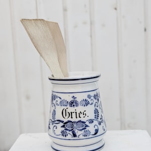 Könnte beinhalten: Weißer Keramiktopf mit blauen Blumenmustern und dem Wort "Gries" in schwarzer Schrift. Zwei Holzutensilien befinden sich im Topf. Der Topf hat einen blauen Rand und Boden, vor weißem Hintergrund.
