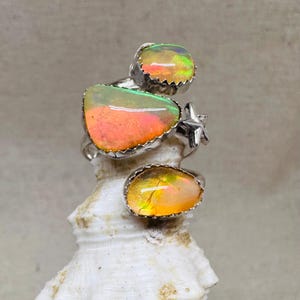 May include: A silver ring featuring three bezel-set, irregular-shaped opals displaying vibrant flashes of orange, yellow, and green. The metal band includes a small, five-pointed star accent positioned between the two larger opal settings.