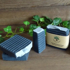 Puede incluir: Tres barras rectangulares de jab&oacute;n de carb&oacute;n activado gris oscuro con textura acanalada. Una barra tiene una etiqueta de papel color canela con un logotipo de &aacute;rbol y el texto The Soap Tree, Handmade in Yorkshire, Activated Charcoal Soap, Tea Tree & Rosemary.
