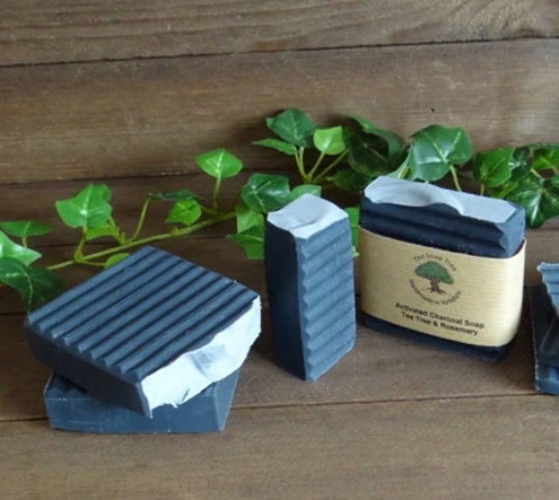 Puede incluir: Tres barras rectangulares de jab&oacute;n de carb&oacute;n activado gris oscuro con textura acanalada. Una barra tiene una etiqueta de papel color canela con un logotipo de &aacute;rbol y el texto The Soap Tree, Handmade in Yorkshire, Activated Charcoal Soap, Tea Tree & Rosemary.