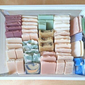 May include: A white wire basket filled with a variety of handmade soap bars. The bars are in different colours and patterns, including green, brown, white, and blue.