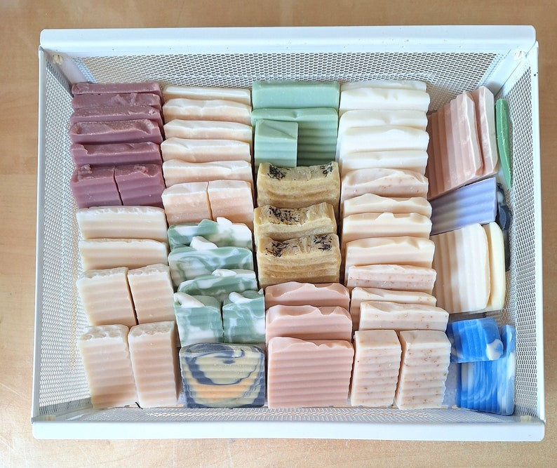 May include: A white wire basket filled with a variety of handmade soap bars. The bars are in different colours and patterns, including green, brown, white, and blue.