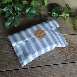 Puede incluir: Un peque&ntilde;o paquete rectangular envuelto en papel de rayas azul claro y blanco, sellado con una pegatina redonda marr&oacute;n que presenta el texto "handmade with love" y un peque&ntilde;o icono de coraz&oacute;n rojo, creando una presentaci&oacute;n sencilla y limpia.