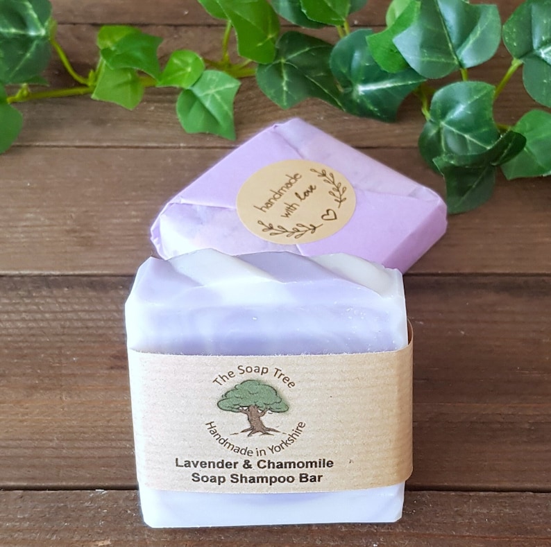 May include: A purple and white soap shampoo bar wrapped in brown paper with the text "The Soap Tree Handmade in Yorkshire Lavender & Chamomile Soap Shampoo Bar". The bar is sitting on a wooden surface.