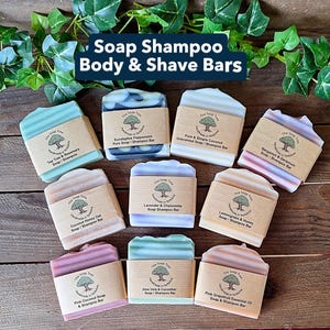 May include: An assortment of handmade soap and shampoo bars in various colors, including green, blue, pink, and beige. Each bar is labeled with its scent and ingredients, such as tea tree, eucalyptus, and coconut. The bars are arranged on a wooden surface.