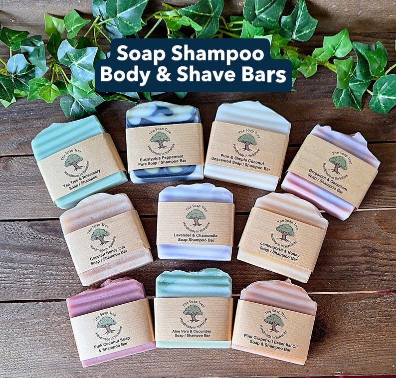 May include: An assortment of handmade soap and shampoo bars in various colors, including green, blue, pink, and beige. Each bar is labeled with its scent and ingredients, such as tea tree, eucalyptus, and coconut. The bars are arranged on a wooden surface.
