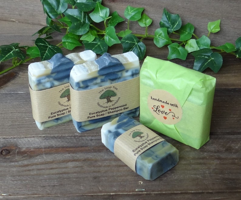 May include: Several bars of handmade soap are displayed, some wrapped in brown paper and others in green tissue. The soap bars have a marbled appearance with blue and white swirls. One bar is wrapped with a label that reads "Eucalyptus Peppermint Pure Soap / Shampoo Bar."