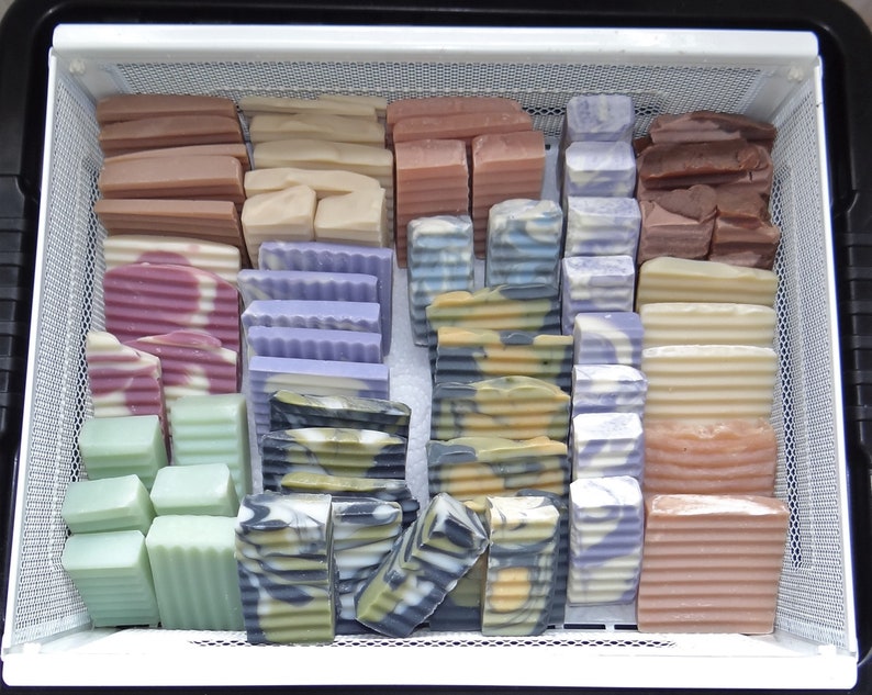 May include: A white plastic container with a wire mesh insert filled with handmade bars of soap in various colours and patterns. The soap bars are arranged in rows and columns.