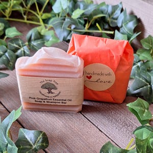 May include: A pink grapefruit essential oil soap and shampoo bar with a light pink top, wrapped in a brown paper band. The band has text that reads "The Soap Tree" and "Handmade in Yorkshire". Beside it is a wrapped package with a sticker that says "handmade with love".