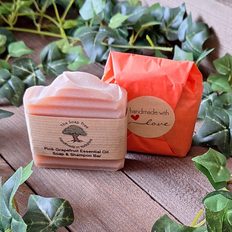 May include: A pink grapefruit essential oil soap and shampoo bar with a light pink top, wrapped in a brown paper band. The band has text that reads "The Soap Tree" and "Handmade in Yorkshire". Beside it is a wrapped package with a sticker that says "handmade with love".