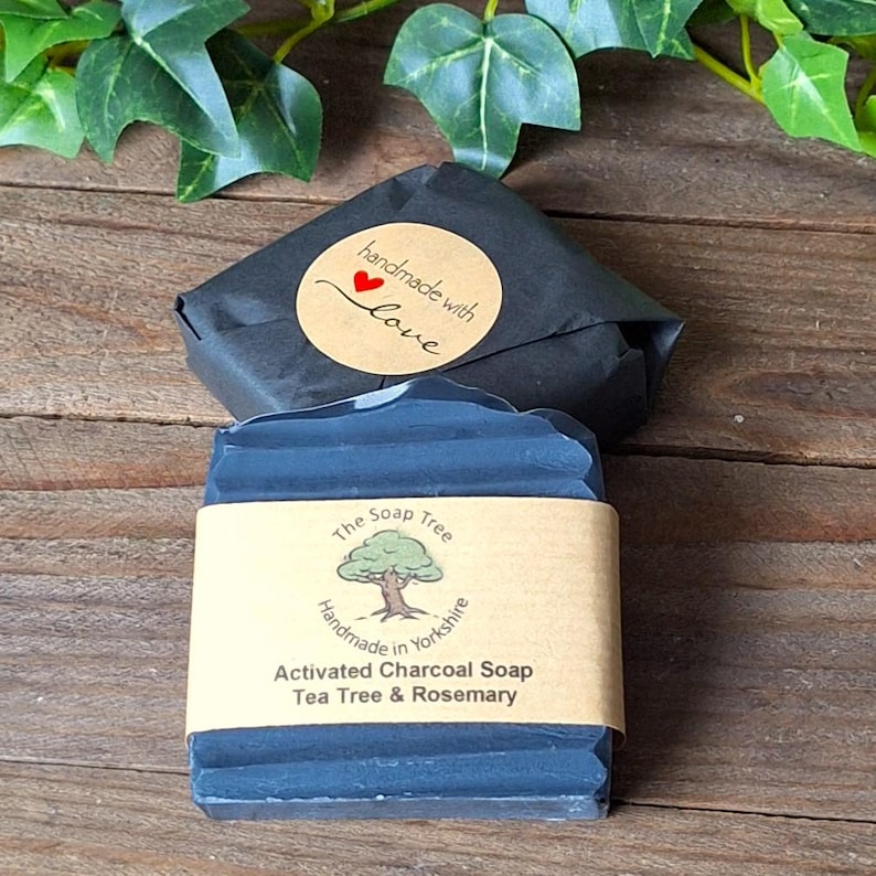 Puede incluir: Una pastilla rectangular de jab&oacute;n de carb&oacute;n activado gris oscuro envuelta en una etiqueta de papel color canela con un logotipo de &aacute;rbol y el texto The Soap Tree Handmade in Yorkshire Activated Charcoal Soap Tea Tree & Rosemary, junto a un jab&oacute;n negro.