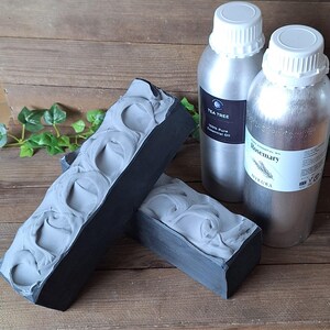 Puede incluir: Dos barras rectangulares de jab&oacute;n gris oscuro con una textura en espiral en la parte superior, colocadas junto a dos botellas de metal plateado con tapas blancas. Una botella tiene la etiqueta Tea Tree y la otra tiene la etiqueta Rosemary.