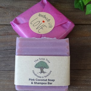 May include: A pink coconut soap and shampoo bar from The Soap Tree, handmade in Yorkshire, England. The bar is wrapped in a pink paper with a sticker that says "handmade with LOVE". The soap is on a wooden surface.