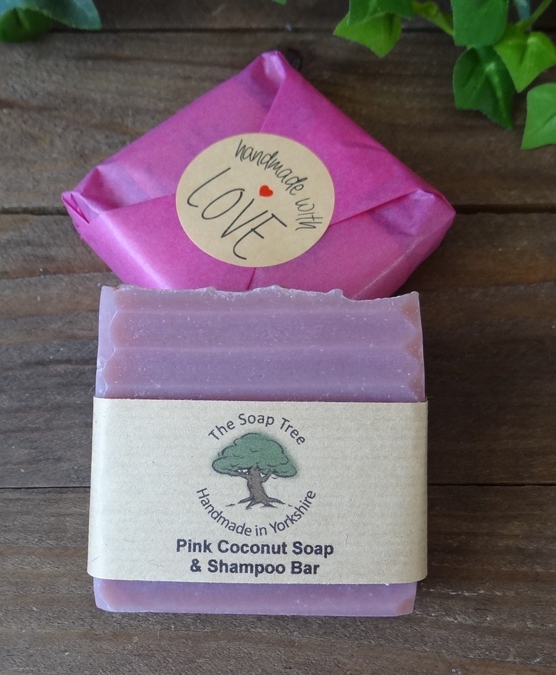 May include: A pink coconut soap and shampoo bar from The Soap Tree, handmade in Yorkshire, England. The bar is wrapped in a pink paper with a sticker that says "handmade with LOVE". The soap is on a wooden surface.